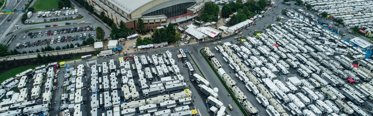 An aerial view of the Hershey RV show, showcasing a sprawling outdoor event with rows of gleaming recreational vehicles (RVs) of various sizes and designs. People can be seen exploring the RVs, and colorful tents and booths line the pathways, creating an exciting atmosphere for RV enthusiasts.